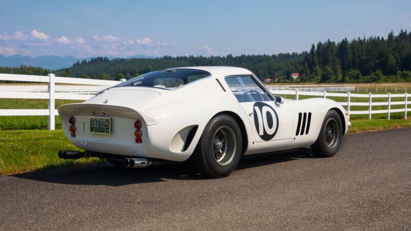 A classic Factory White Ferrari 250 GTO with the number 10 on the door is parked on an asphalt road next to a white fence and grassy fields, with trees and mountains in the background.