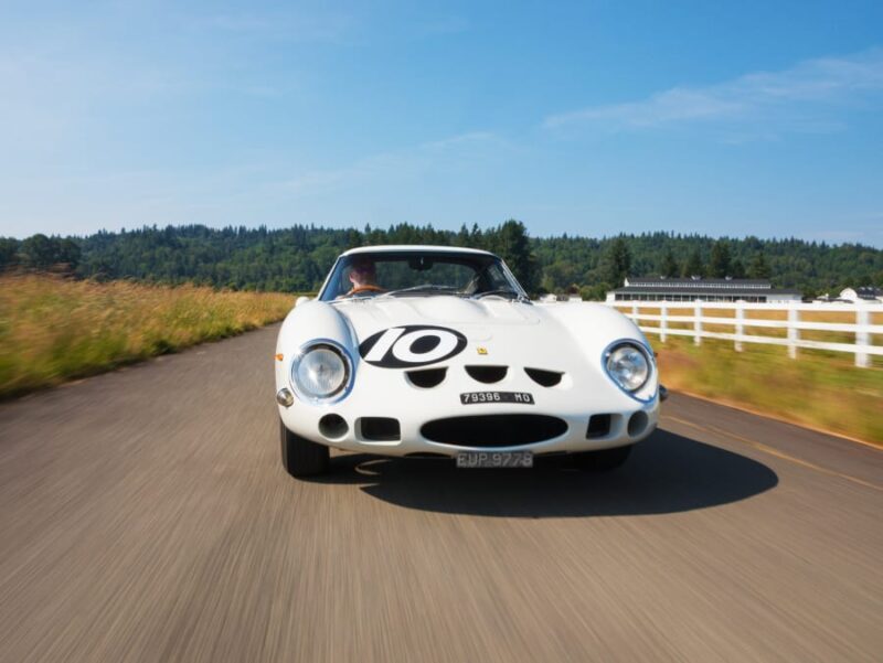 A classic white Ferrari 250 GTO with the number 10 on the hood drives on a country road, passing a white fence, grass, and trees under a clear blue sky—ready for its moment at the Kissimmee 2026 Mecum auction.