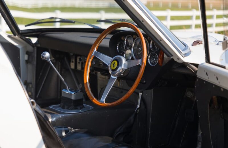 Close-up view of the interior of a Factory White Ferrari 250 GTO, showcasing its wooden steering wheel with Ferrari logo, dashboard gauges, and manual gear shift lever—an iconic sight at the Mecum Kissimmee Auction.