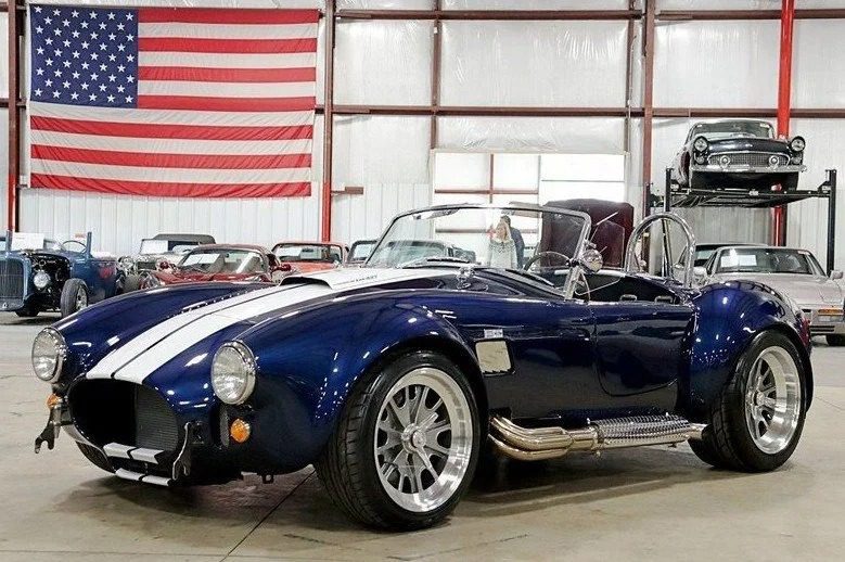 A blue Shelby Cobra with white racing stripes is parked indoors among other classic muscle cars, with an American flag hanging on the wall in the background.