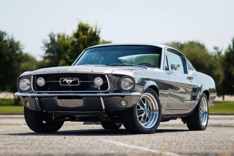 A classic silver Ford Mustang coupe, a standout among Classic Muscle Cars, is parked outdoors on asphalt, with trees and a clear sky in the background.