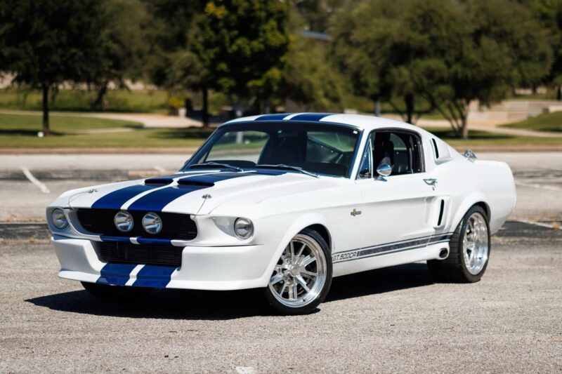 A white classic Ford Mustang with blue racing stripes is parked on an empty asphalt lot, showcasing the timeless appeal of classic muscle cars amidst a background of trees and grass.