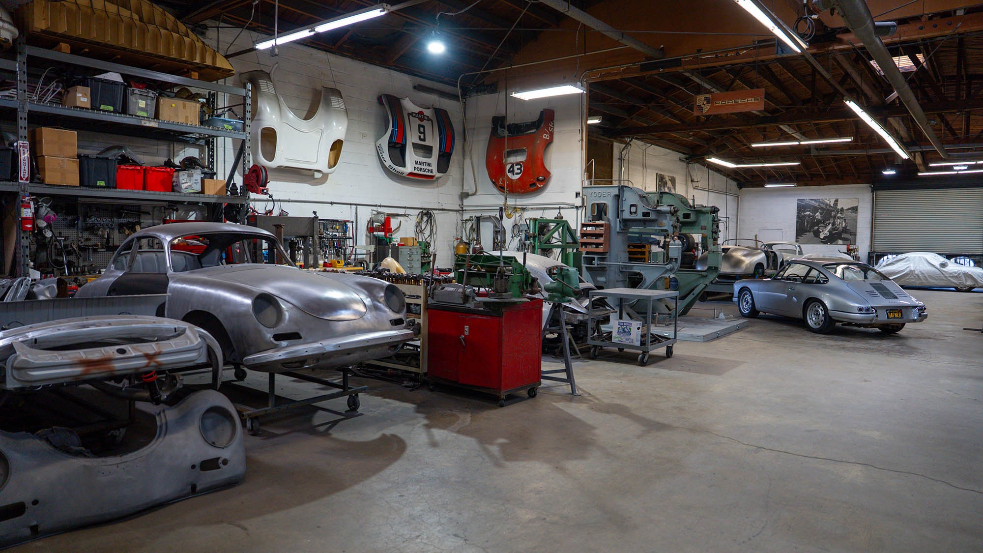 A spacious auto workshop with classic cars under restoration, including a Prototype Porsche 912 and other vintage models, tools, car parts, hanging car hoods on the walls, and a silver car—possibly a 1968 Porsche—on the left.