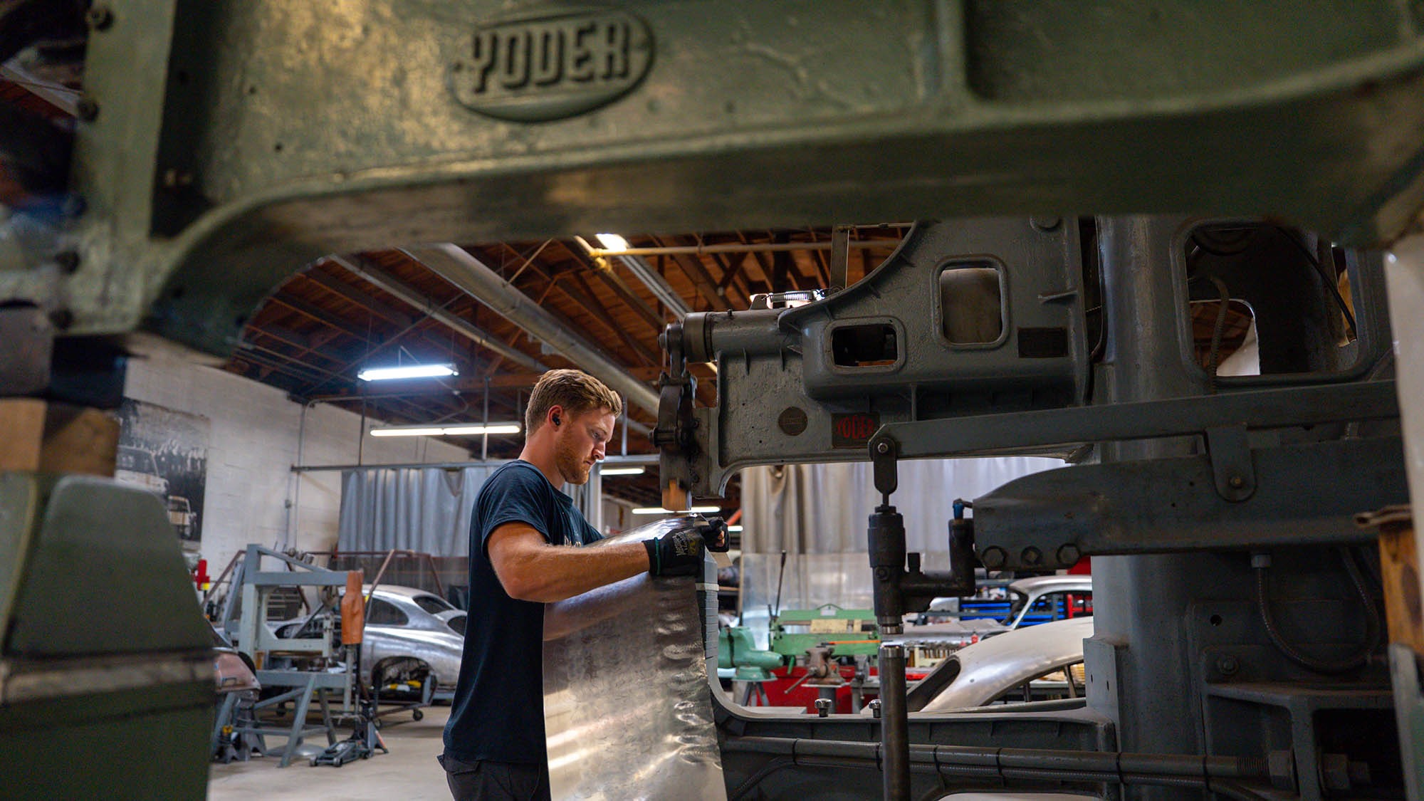 A man in a workshop uses a large Yoder metal forming machine to shape a sheet of metal, with various tools and Porsche 912 parts from a 1968 Prototype visible in the background, evoking a scene worthy of Petrolicious.