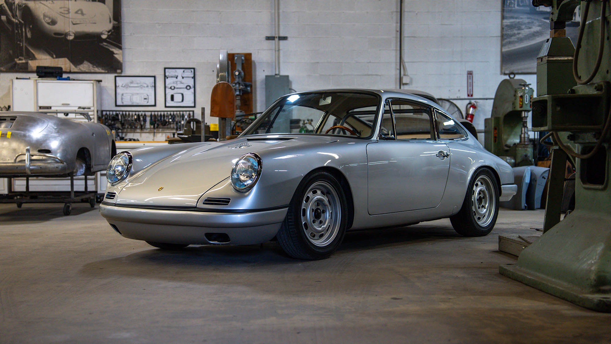 A silver classic Porsche 912 coupe, reminiscent of a Petrolicious feature, is parked inside a workshop, surrounded by automotive tools, equipment, and car parts.