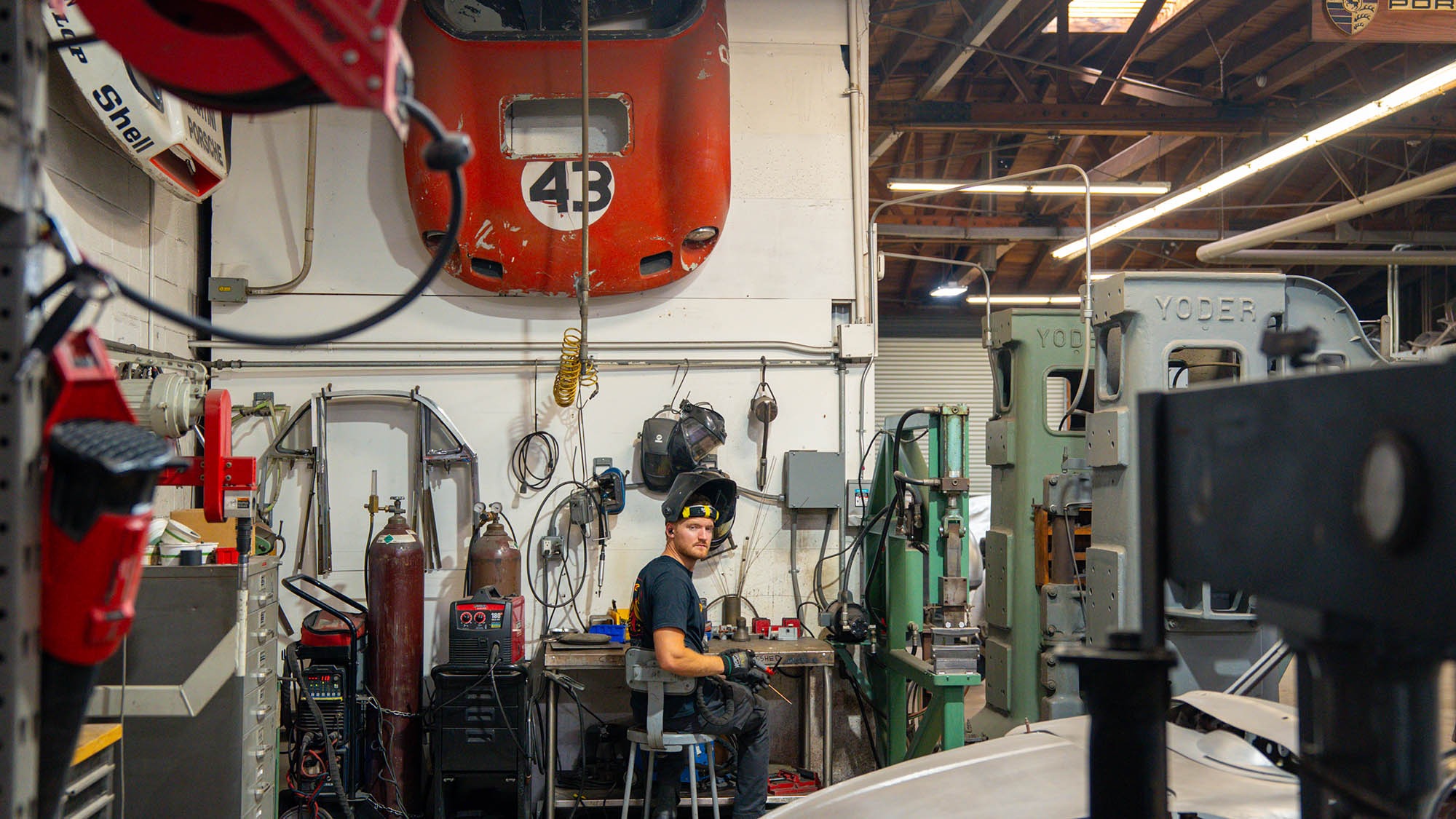A man wearing safety goggles sits on a stool in an industrial workshop, surrounded by machinery and tools, with a red Porsche 912 hood mounted on the wall above him—a setting worthy of a Petrolicious feature.