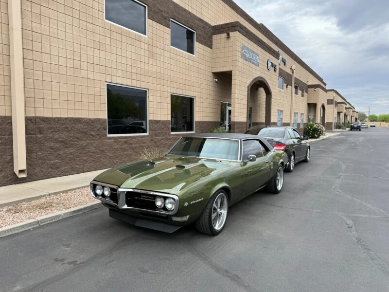 A green classic Pontiac Firebird, a standout among Classic Muscle Cars, is parked in front of a modern beige office building, with another car behind it on an overcast day.