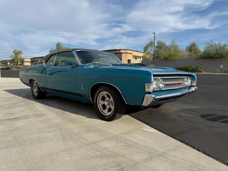 A blue classic Ford Torino GT, one of the iconic classic muscle cars, is parked on a driveway near a curb, with modern buildings and trees in the background under a partly cloudy sky.