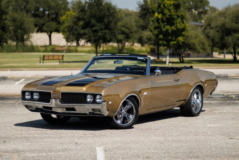 A gold and black vintage convertible, reminiscent of classic muscle cars, is parked in an empty lot with grass and trees in the background.