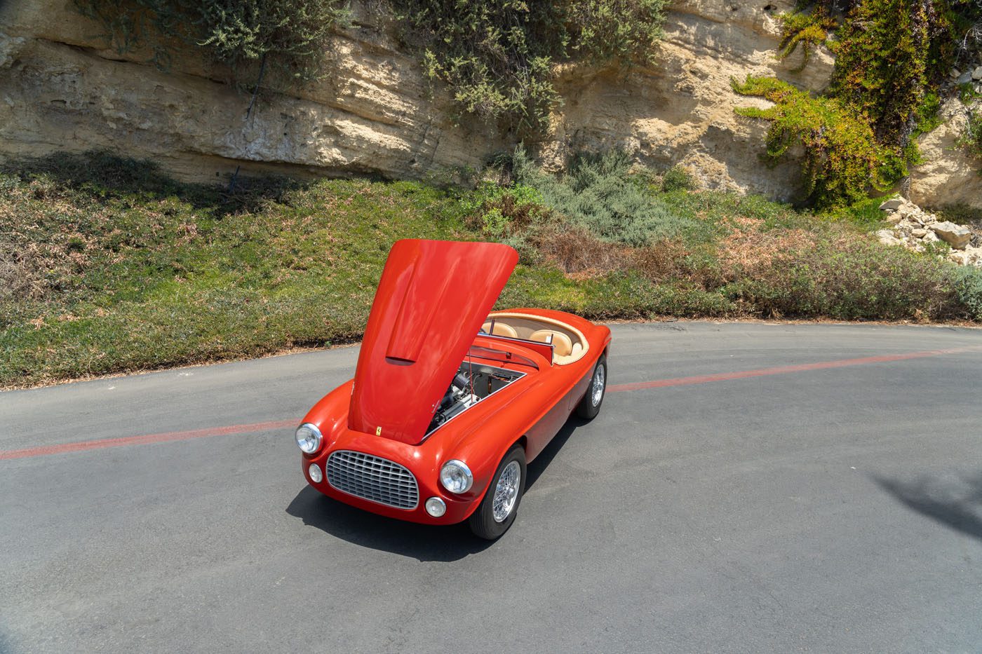 A classic red convertible car with its hood open is parked on a curved road beside a rocky, grassy hillside, reminiscent of the Ferrari Legends at the upcoming Monterey 2025 Mecum Auction.