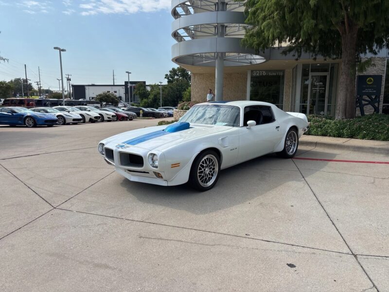 A white classic muscle car with blue racing stripes is parked outside a dealership, showcasing classic muscle cars alongside several modern high-performance cars in the background.