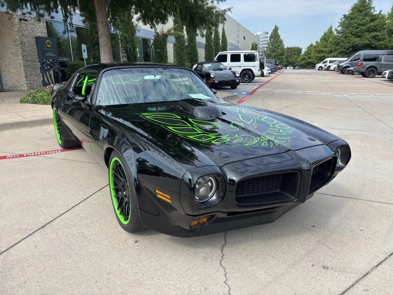 A black classic muscle car with green-accented wheels and a green firebird decal on the hood is parked outside a modern building—a true icon for Muscle Cars enthusiasts.