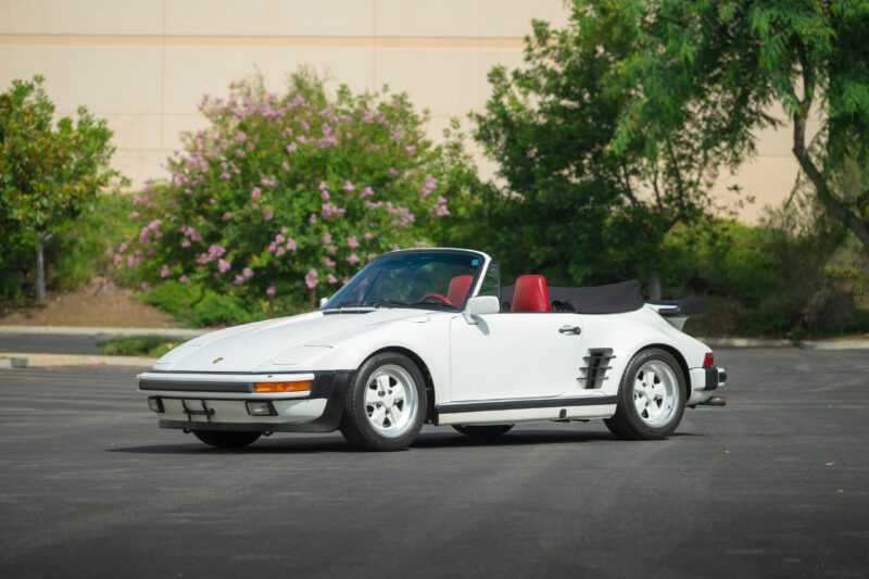 A white convertible sports car with a red interior is parked on an asphalt surface, framed by trees and flowering bushes—a standout sight worthy of the 2025 Bonhams Monterey: 10 Must-See Cars Crossing the Block at The Quail.
