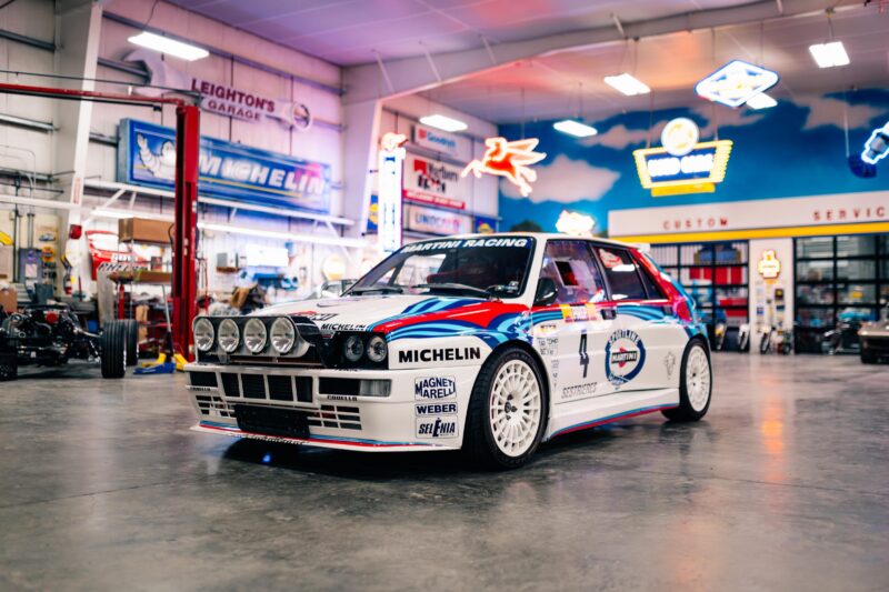 A vintage rally car with iconic Martini Racing livery is parked inside a well-lit garage, decorated with neon signs and automotive memorabilia—a scene any enthusiast at the 2025 Bonhams Monterey: 10 Must-See Cars Crossing the Block at The Quail would admire.