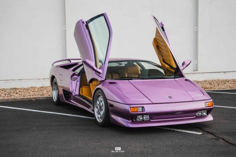 A purple Lamborghini Diablo with scissor doors open, parked on a blacktop surface near a white wall—a true icon of 90s cars for sale, radiating nostalgia and automotive history.