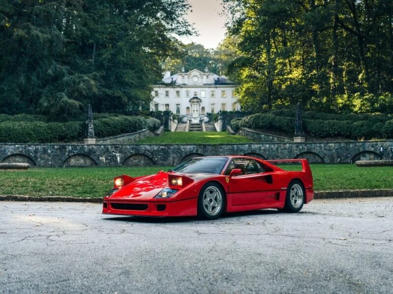 A red sports car with raised headlights, radiating nostalgia, is parked on a driveway in front of a large, white mansion surrounded by trees and greenery.