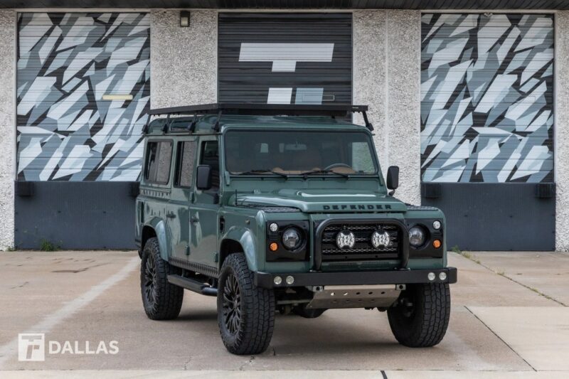 A green Land Rover Defender, evoking nostalgia and history, is parked on a concrete surface in front of a building with geometric designs on the garage doors.