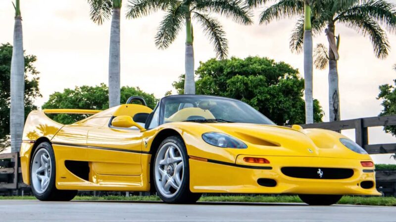 A yellow Ferrari convertible sports car, featured among the Top 10 Results and Highlights at the 2025 RM Sotheby's Monterey Auction, is parked on a paved surface with palm trees and greenery in the background.
