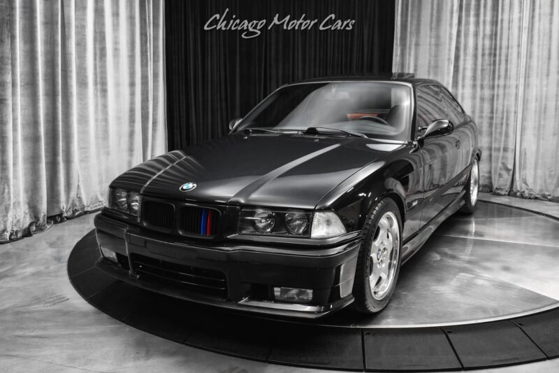 A black BMW coupe is displayed on a rotating platform in a showroom with a "Chicago Motor Cars" sign above, evoking nostalgia for classic 90s cars for sale and celebrating the brand's rich history.
