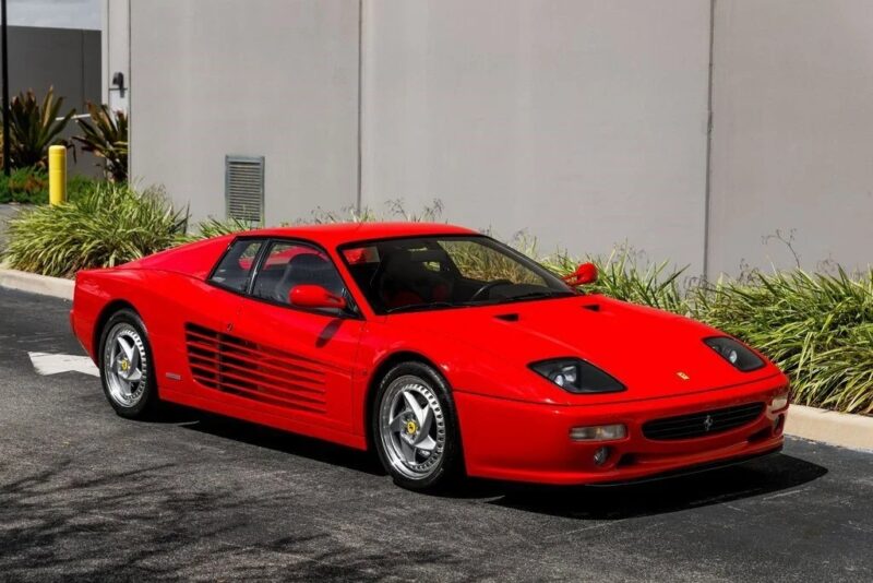 A red Ferrari sports car, echoing the nostalgia of 90s cars for sale, is parked on a paved road next to a gray building and lush green landscaping.
