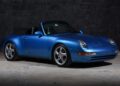 A high-performance blue Porsche 911 convertible is parked indoors on a concrete floor with a dark, textured wall in the background.