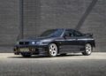 A black 1996 Nissan Skyline GT-R R33 sports car is parked on a concrete surface in front of a gray brick wall.