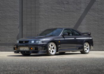 A black 1996 Nissan Skyline GT-R R33 sports car is parked on a concrete surface in front of a gray brick wall.
