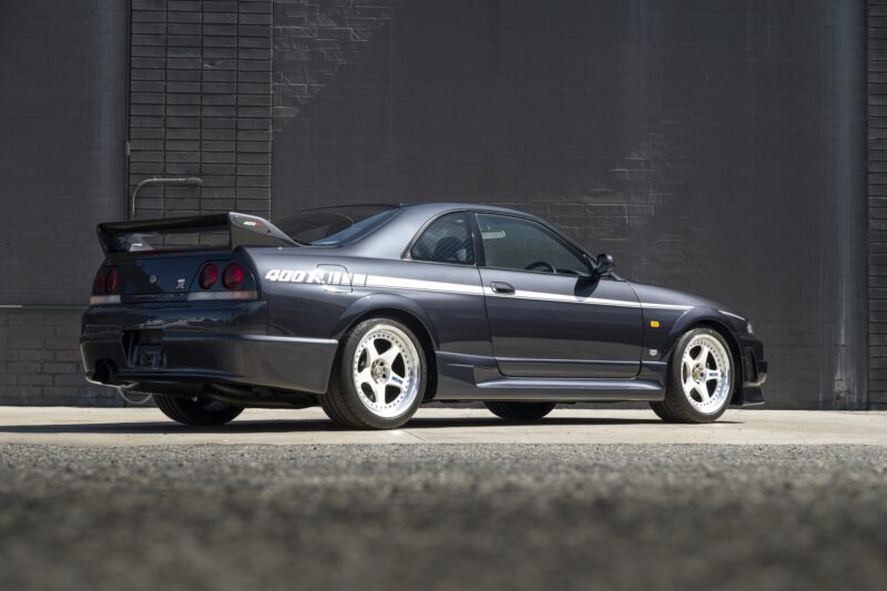 A dark gray 1996 Nissan Skyline GT-R R33 coupe is parked on pavement in front of a brick wall, viewed from the rear passenger side, resembling the legendary NISMO 400R.