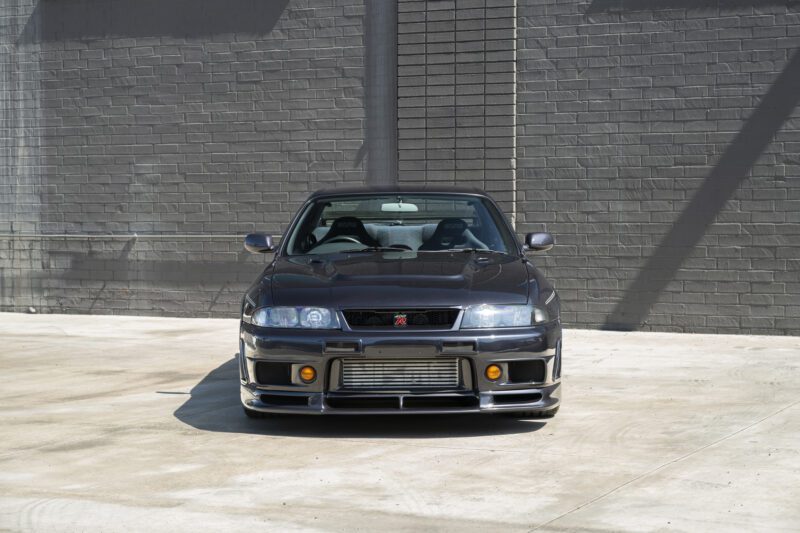 Front view of a dark grey 1996 Nissan Skyline GT-R parked on a concrete surface in front of a gray brick wall, inspired by the iconic NISMO 400R.