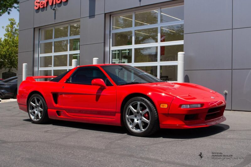A red Acura NSX sports car—an icon among 90s cars for sale—is parked in front of a grey building with large windows and a "Service" sign above, evoking a sense of nostalgia.
