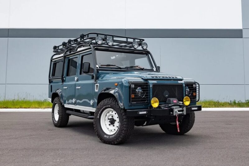 A blue Land Rover Defender with roof rack, spotlights, and off-road accessories—pure 90s cars nostalgia—is parked on an asphalt surface in front of a light grey building.