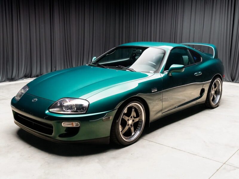 A green sports car with a rear spoiler is parked indoors on a concrete floor, dark curtains in the background—evoking nostalgia for 90s cars for sale and celebrating automotive history.