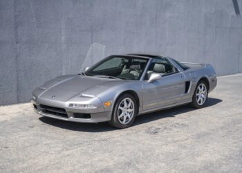 A silver two-door sports car, reminiscent of 90s cars for sale, is parked on a concrete surface next to a gray textured wall.