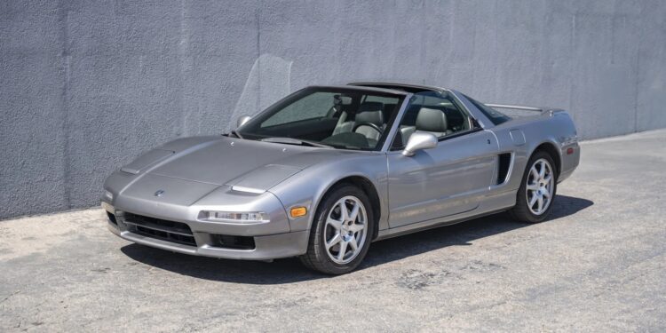 A silver two-door sports car, reminiscent of 90s cars for sale, is parked on a concrete surface next to a gray textured wall.