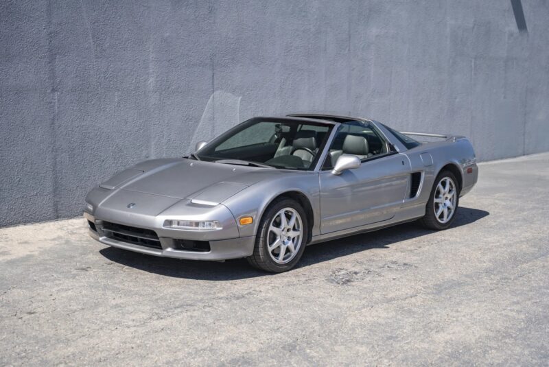 A silver two-door sports car, reminiscent of 90s cars for sale, is parked on a concrete surface next to a gray textured wall.