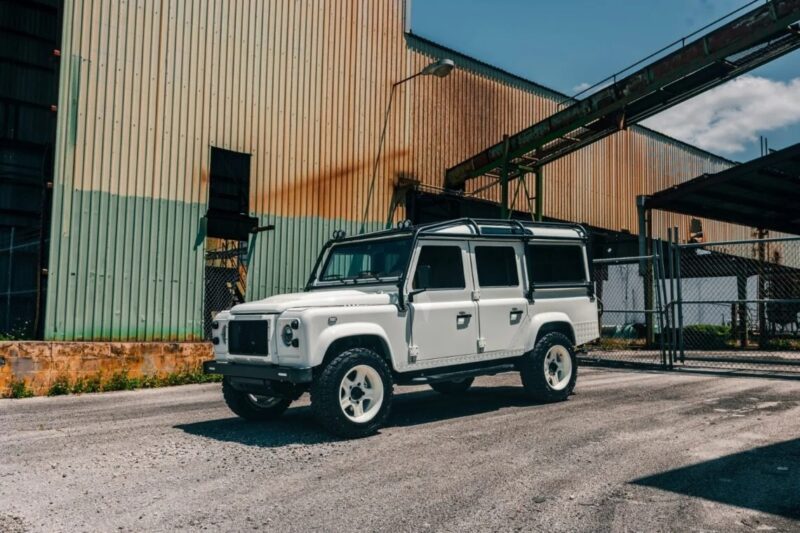 A white SUV with off-road features is parked on a paved surface near an industrial building with metal siding and rust stains, evoking nostalgia for the rugged 90s cars for sale in automotive history.