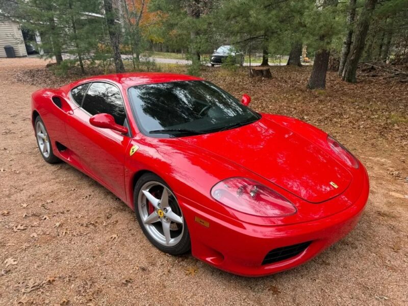A red Ferrari sports car, evoking nostalgia, is parked on a dirt driveway surrounded by trees and fallen leaves.