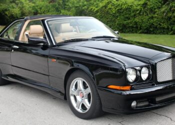 A black Bentley coupe with tan interior parked on a paved surface, featuring chrome wheels and a distinctive mesh grille, evokes nostalgia with its classic design, set against green foliage in the background.