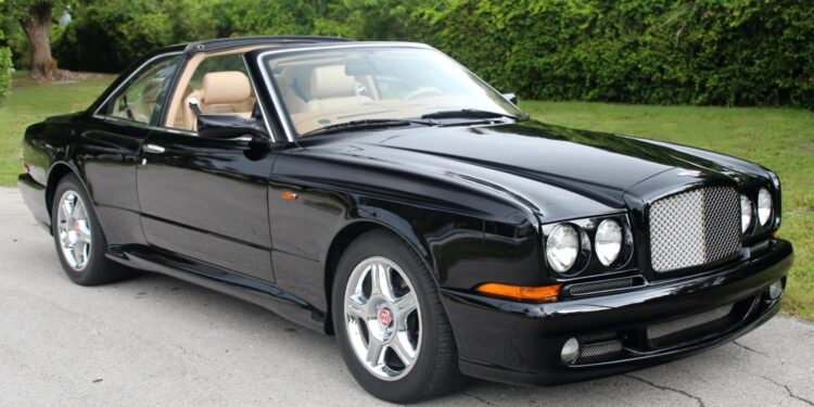 A black Bentley coupe with tan interior parked on a paved surface, featuring chrome wheels and a distinctive mesh grille, evokes nostalgia with its classic design, set against green foliage in the background.