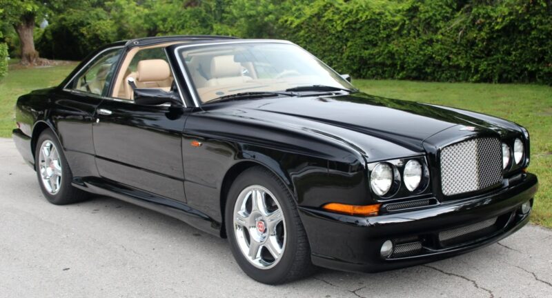 A black Bentley coupe with tan interior parked on a paved surface, featuring chrome wheels and a distinctive mesh grille, evokes nostalgia with its classic design, set against green foliage in the background.