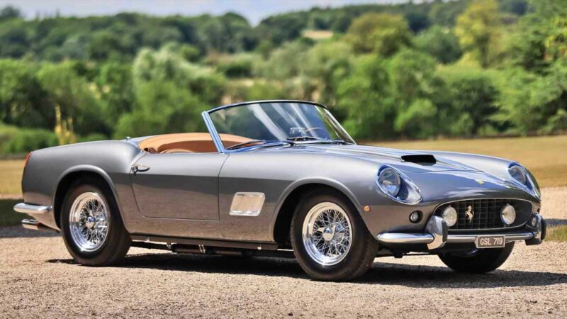 A silver classic convertible sports car with tan interior is parked outdoors on a gravel surface with greenery in the background, potentially one of the 10 Most Expensive Cars To Sell At 2025 Monterey Car Week Auctions.
