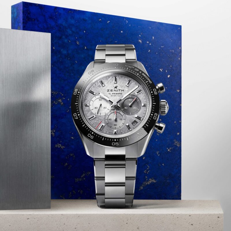 Stainless steel Zenith Chronomaster Sport chronograph wristwatch with a silver dial, black bezel, and metal bracelet displayed against blue and gray textured backgrounds.