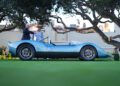 A light blue vintage sports car is parked on grass, with two men standing nearby under large trees, and a building in the background.
