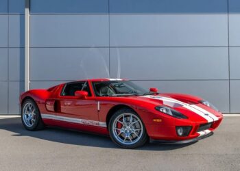 A red Ford GT sports car with white racing stripes, one of the best 2000s cars, is parked on a paved surface in front of a gray building.