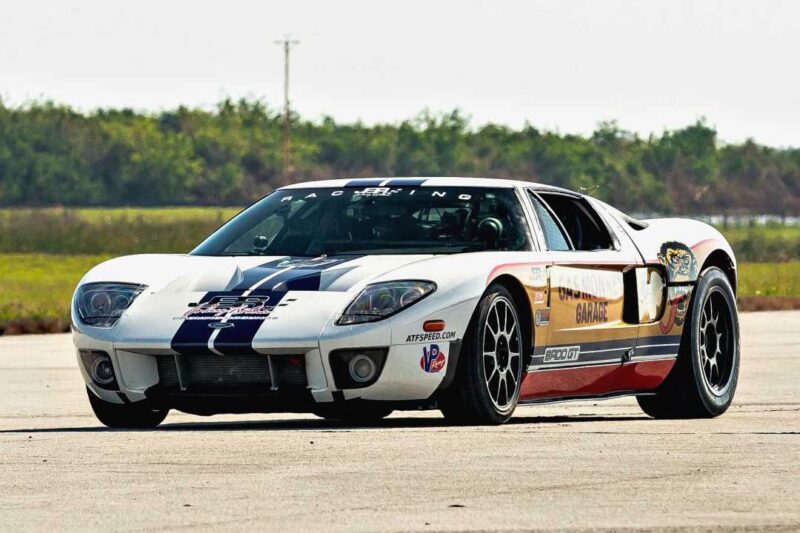 A white sports car with blue racing stripes, sponsor decals, and black wheels is parked on a paved surface with trees in the background—reminiscent of the 330 MPH Dream: Jonny Bohmer’s BADD GT aiming for a historic run in 2026.