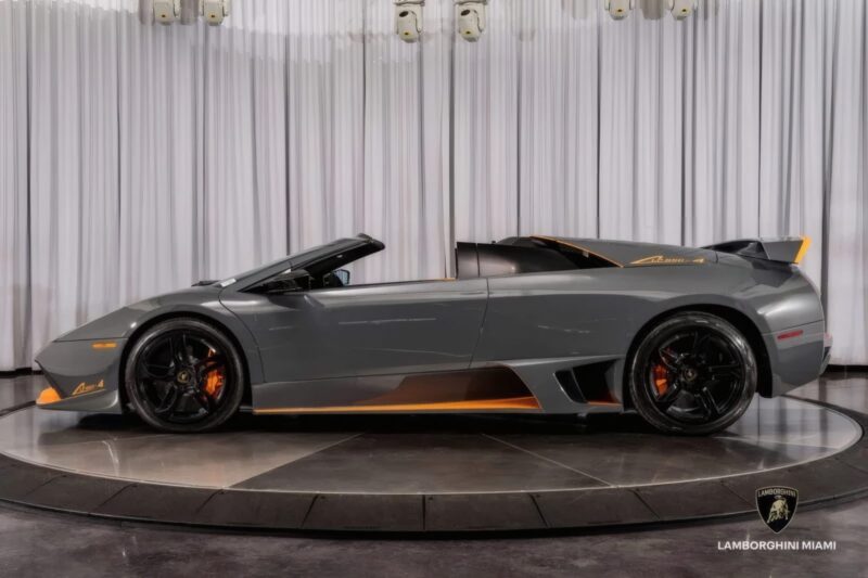 A grey Lamborghini convertible sports car with black wheels and orange accents is displayed indoors on a rotating platform—the #50-of-50: LAST Lamborghini Murcielago LP 650-4 Roadster for Sale at Prestige Imports.
