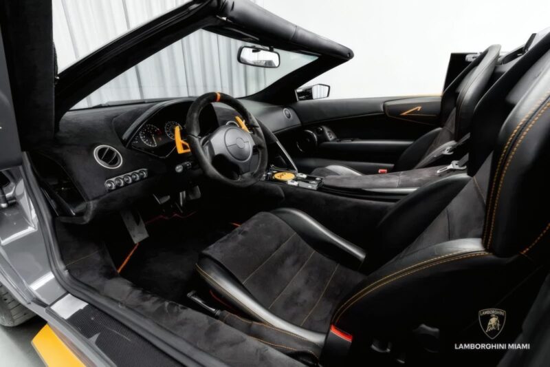 Interior view of a Lamborghini convertible showing black leather seats with yellow stitching, dashboard controls, and the Lamborghini Miami logo in the corner. #50-of-50: LAST Lamborghini Murcielago LP 650-4 Roadster for Sale at Prestige Imports.