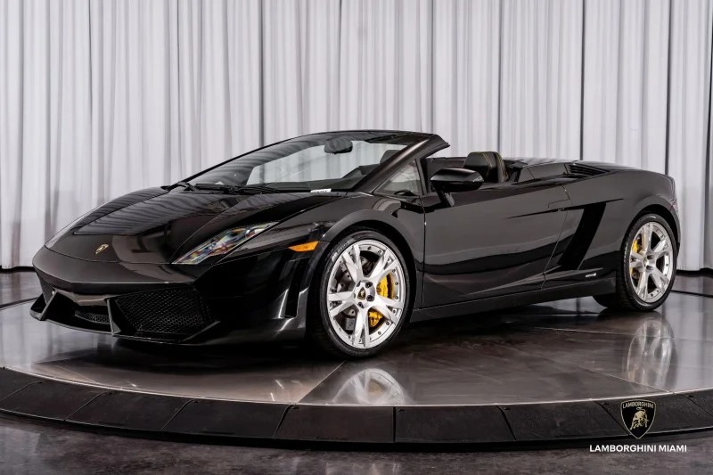 A Lamborghini Gallardo V10 supercar convertible is displayed on a circular platform with silver wheels, yellow brake calipers, and a white curtain backdrop.