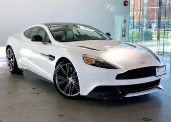 A white Aston Martin coupe, one of the finest exotic cars, is parked indoors near large glass windows, with sunlight reflecting off its body and black alloy wheels.