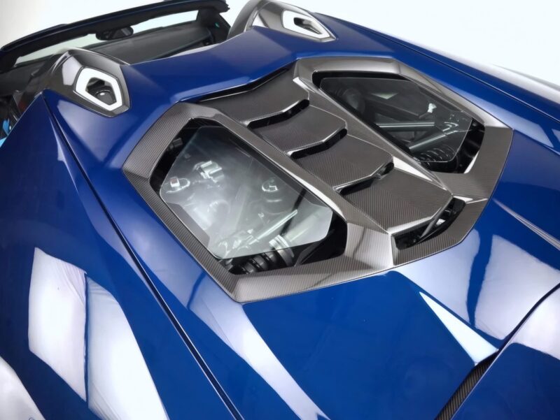 Close-up of a blue Lamborghini Centenario Roadster's rear engine cover—only 10 in the U.S.—featuring carbon fiber accents and two transparent panels revealing parts of the engine.